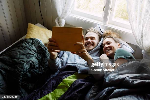 high angle view of smiling friends watching movie over digital tablet while lying on bed in cottage - zusehen stock-fotos und bilder