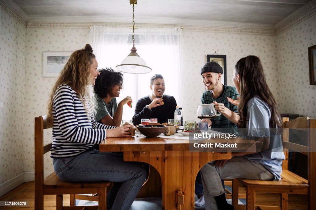 Happy friends talking while enjoying food and drink on table at home