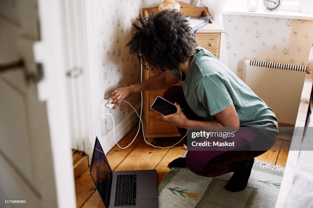 Side view of young woman plugging mobile phone charger in electrical outlet at home seen through doorway
