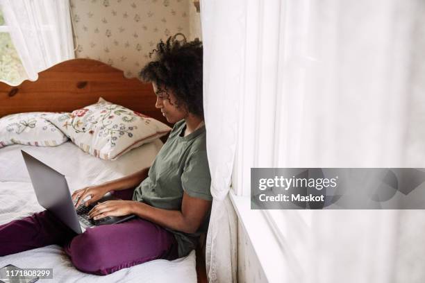 side view of young woman with curly hair using laptop while sitting on bed at home - pillow icon stock pictures, royalty-free photos & images