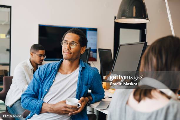 thoughtful male computer programmer looking away while sitting by colleagues at desk in office - african american man day dreaming stock pictures, royalty-free photos & images