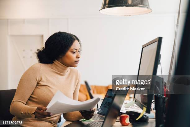 focused female it professional holding document while looking at computer in creative office - black woman computer programmer stock pictures, royalty-free photos & images