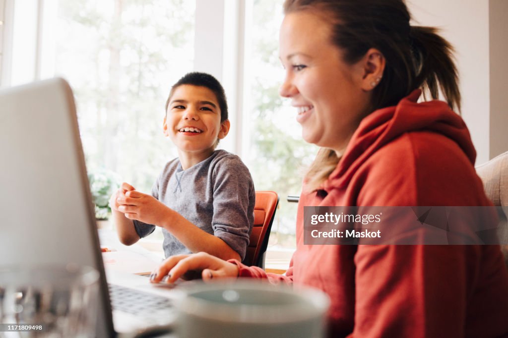 Mother using laptop while sitting with happy autistic son in living room at home