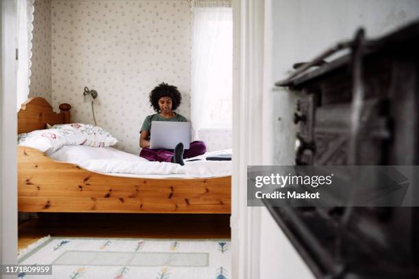 young woman using laptop while relaxing on bed at home seen through doorway - pillow icon stock pictures, royalty-free photos & images