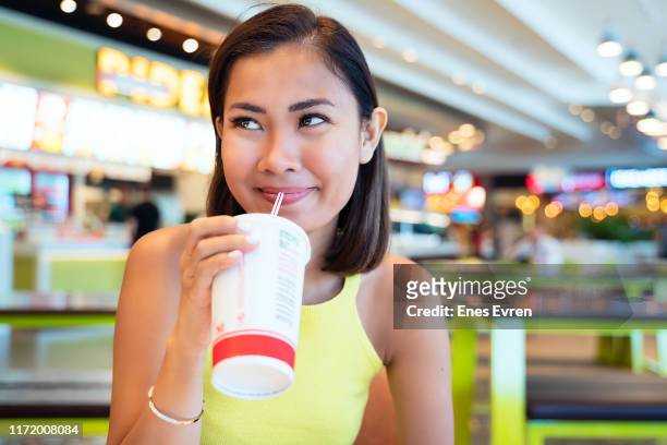 asian woman having fast food, drinking with disposable cup in shopping mall food court - food court stock pictures, royalty-free photos & images