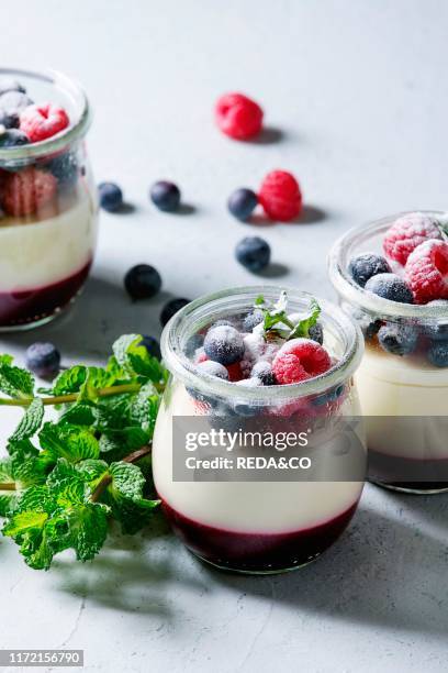 Homemade classic dessert Panna cotta with raspberry and blueberry berries and jelly in jars, decorated by mint and sugar powder over white blue table.