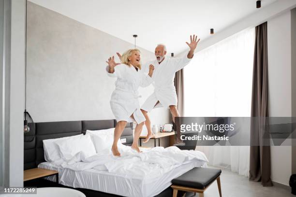 senior couple having fun in hotel room jumping on a bed - couple playful bedroom stock-fotos und bilder