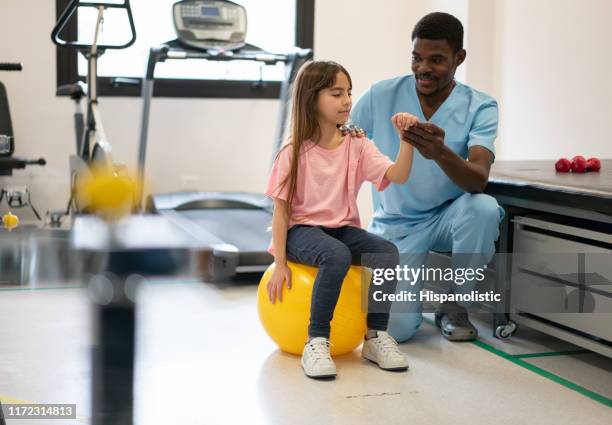 cute little girl sitting on fitness ball while black male therapist holds her arm during physical therapy - orthopedic equipment stock pictures, royalty-free photos & images