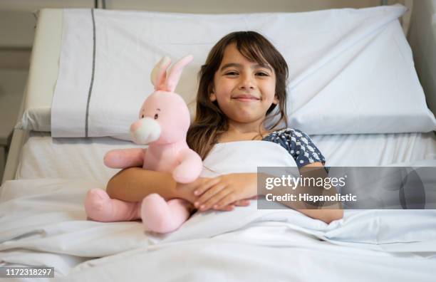 portrait of beautiful little girl hugging her pink rabbit while lying down on hospital bed smiling at camera - girl in hospital bed sick stock pictures, royalty-free photos & images