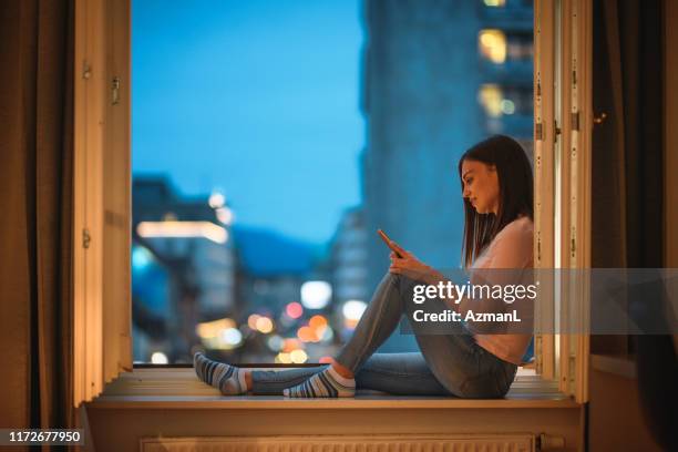 ung kvinna koppla av med smart telefon i öppet fönster i skymningen - open city bildbanksfoton och bilder