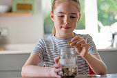 Girl Putting Coins Into Glass Jar Labelled Savings At Home
