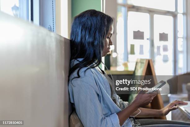 woman at café gebruikt oordopjes en schuift op een smartphone - african american restaurant texting stockfoto's en -beelden