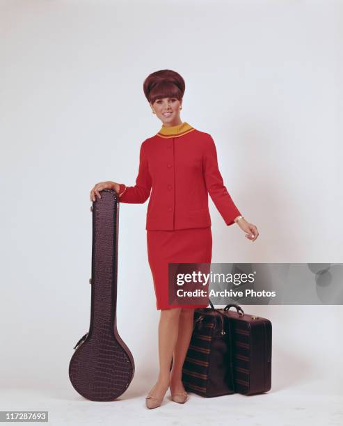 Woman modelling women's fashions in a studio portrait, wearing a red two-piece outfit, with a yellow collar, posing beside two suitcases and a banjo...