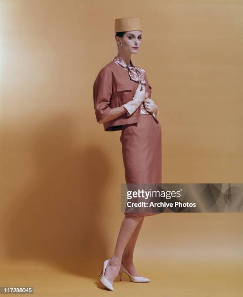 Woman modelling women's fashions in a studio portrait, standing wearing a salmon pink two-piece outfit, with a patterned blouse, white gloves, white...