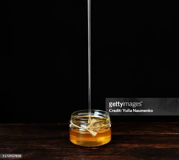 jet of honey pouring into a glass jar, dripping liquid honey, tasty golden yellow. brown wooden table against black background. copyspace. - pot of gold stock-fotos und bilder