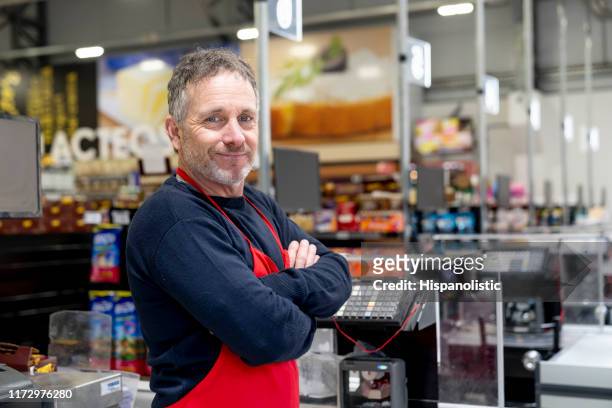 portrait of adult male cashier at the supermarket facing camera smiling with arms crossed - supermarket cashier stock pictures, royalty-free photos & images