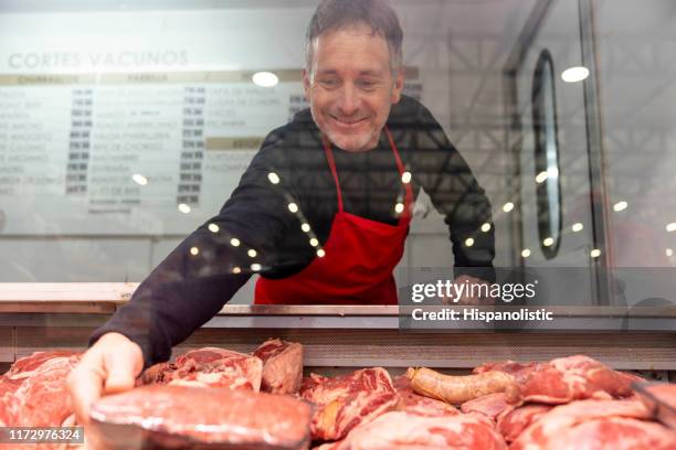 macellaio amichevole che afferra un pezzo di carne da tagliare per un cliente - macellaio foto e immagini stock