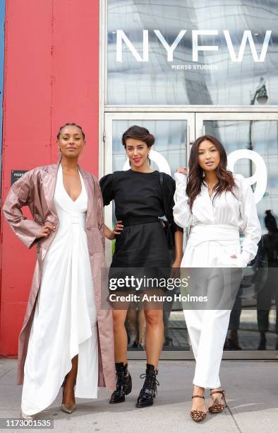 Melanie Liburd is seen wearing a white maxi dress, a mauve trench coat, and metallic pumps and Jeannie Mai is seen wearing white jumpsuit with a...