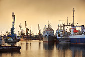 Fishing vessels and docks cranes in the port on the background of a sunset.