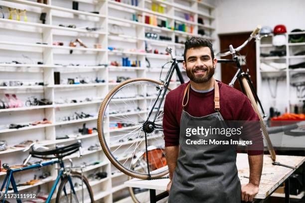 smiling young bearded owner leaning on workbench - bike shop stock pictures, royalty-free photos & images