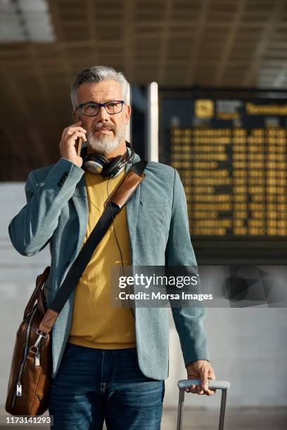 mature man with luggage talking on smart phone - departure board front on fotografías e imágenes de stock
