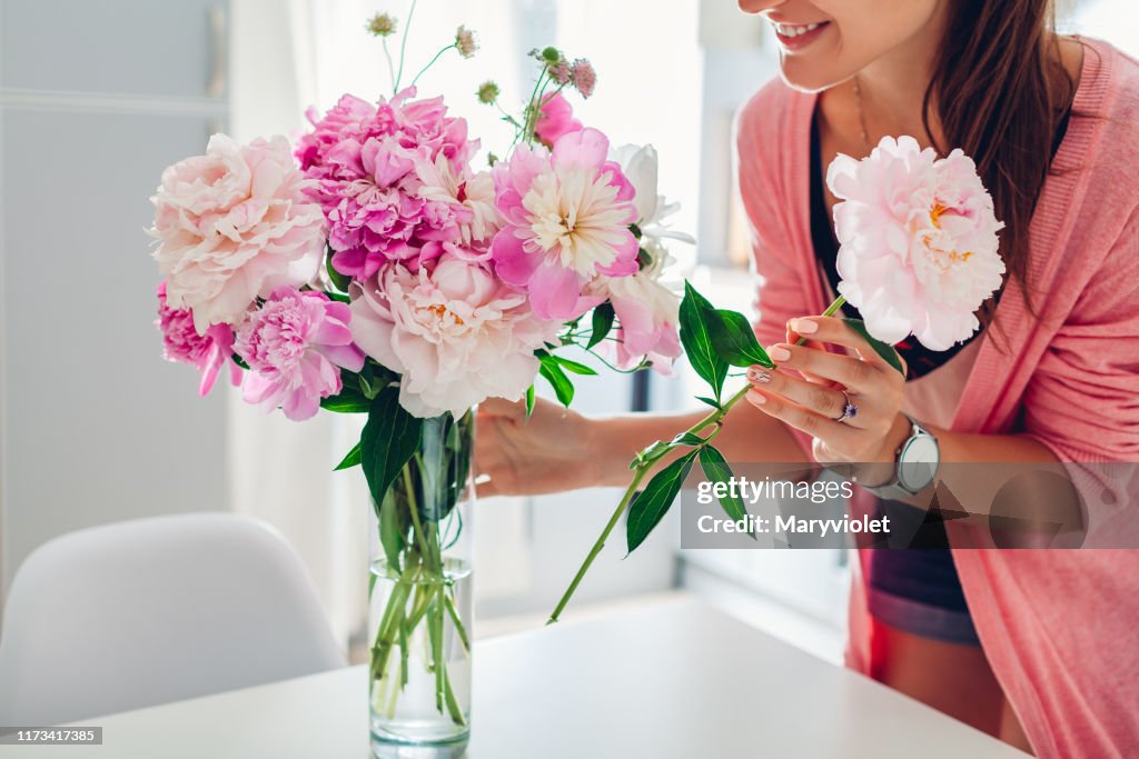 A mulher põr flores dos peônias no vaso. Dona de casa que toma do aconchego e da decoração na cozinha. Compondo bouquet.