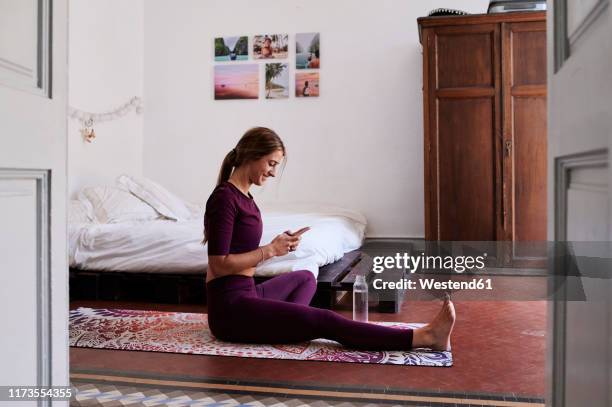 young brunette woman during yoga, using smartphone in student dorm - brunette woman bedroom stock-fotos und bilder
