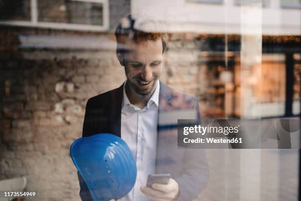 smiling businessman with hard hat looking at cell phone - person in suit construction stock pictures, royalty-free photos & images