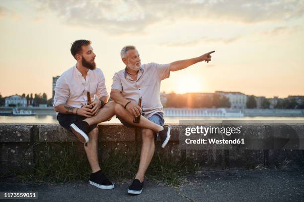 father and adult son sitting on a wall at the riverside at sunset drinking a beer - beer flowing stock pictures, royalty-free photos & images