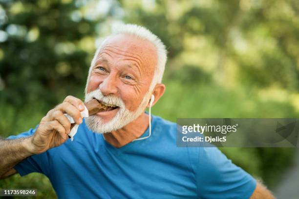 sporty senior man eating a chocolate bar after outdoors training. - senior people training imagens e fotografias de stock