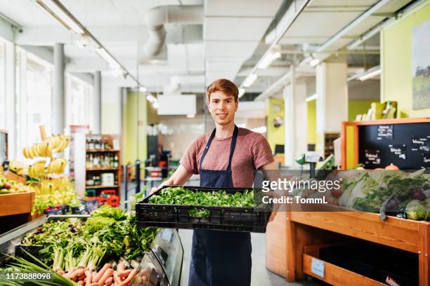 verticale du jeune commis de supermarché retenant la caisse des légumes - shop seller photos et images de collection