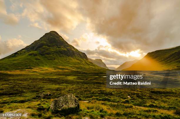 glencoe valley during golden hour sunset in a partly cloudy summer day, sunbeam through the clouds. green grass everywhere. mountains in the background - schottisches hochland stock-fotos und bilder