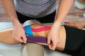 Hands of a female physiotherapist taping light blue medical tape over another pink tape on a patient's knee.