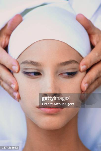 Vogue, October 1985 - Head shot of model Christy Turlington in a white head wrap with another person's hands at her cheekbones and temples, tugging...
