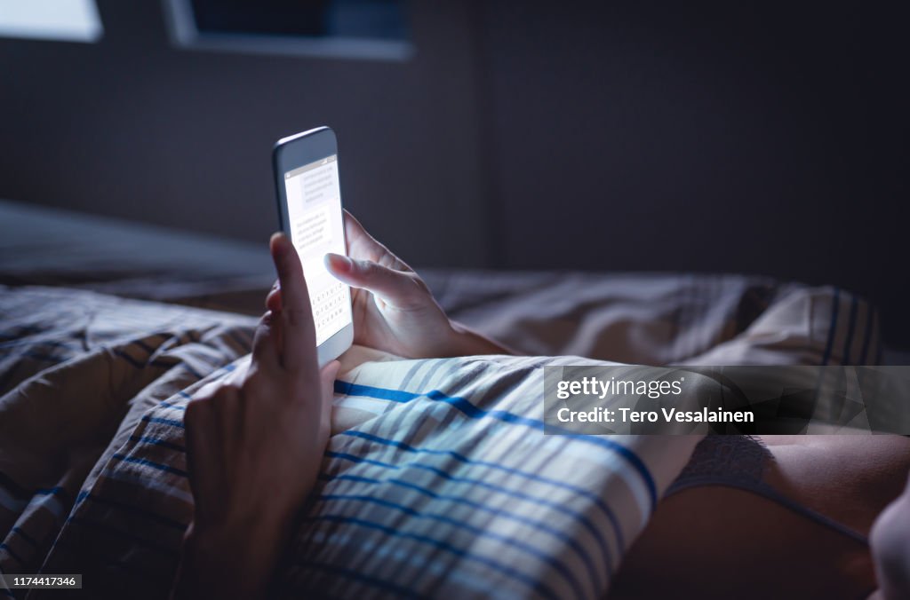 Woman using phone late at night in bed. Person looking at text messages with cell in dark home. Hipster online dating or texting with smartphone. Sexting or cheating concept.