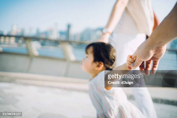 cute little toddler girl holding her mother and father's hand strolling along the promenade against urban cityscape on a lovely sunny day - couple holding hands fotografías e imágenes de stock