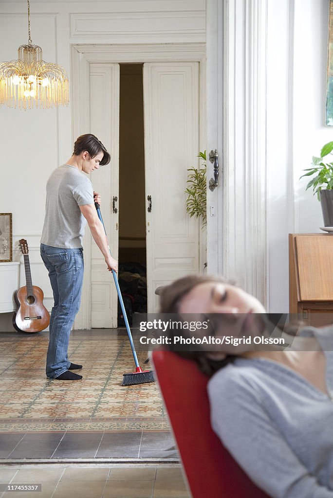 Man sweeping floor at home, woman napping in foreground