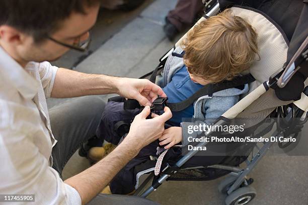 father strapping toddler boy into stroller - stroller stock pictures, royalty-free photos & images