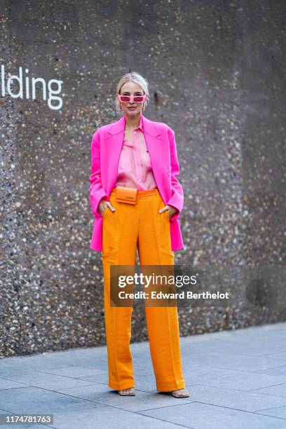 Leonie Hanne wears pink sunglasses, earrings, a pink blazer jacket, a pink shirt, orange flare pants, an orange Jacquemus belt bag, during London...