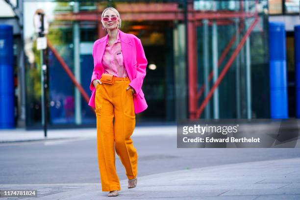 Leonie Hanne wears pink sunglasses, earrings, a pink blazer jacket, a pink shirt, orange flare pants, an orange Jacquemus belt bag, during London...