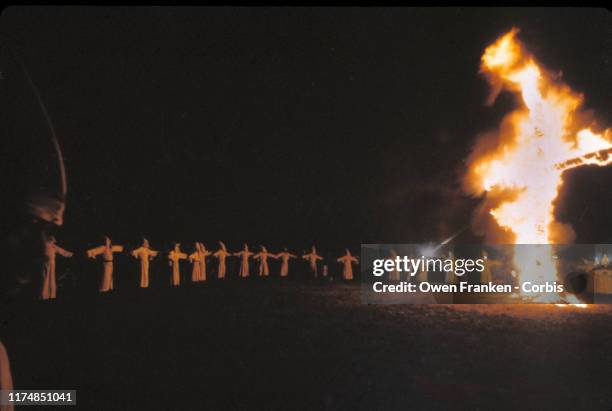 Ku Klux Klan members, in white robes and hoods, gather around a burning cross during a nighttime rally, Scotland, Connecticut, 1970s.