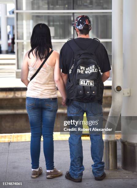 Young couple holds hands as they wait for a train at Union Station in Denver, Colorado. The man is wearing a 'The Elite' brand backback.