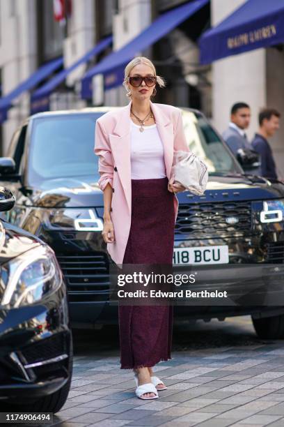 Leonie Hanne wears sunglasses, a pink oversized blazer jacket, a necklace, a white top, a purple wool skirt, white sandals, a bag, during London...