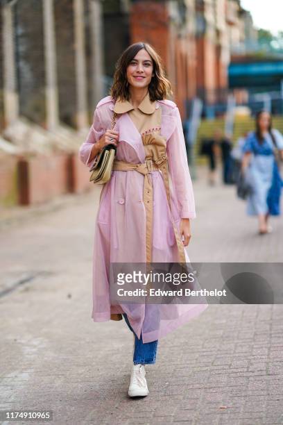 Alexa Chung wears a pale pink transparent trench coat with a beige part, a belt, a bag, blue jeans, white sneakers, during London Fashion Week...