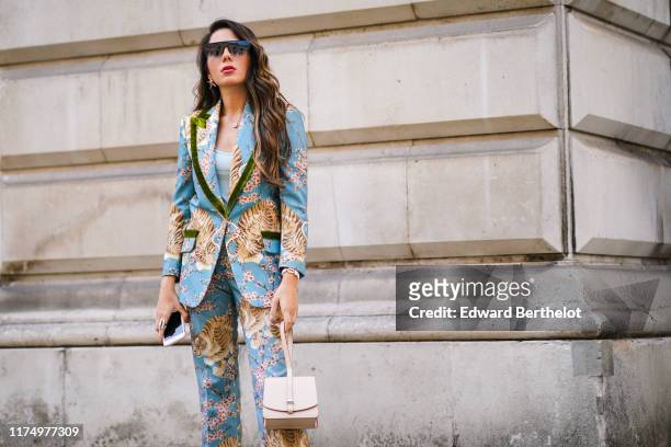 Nurce Erben wears sunglasses, a blazer jacket with floral and tiger print, flare pants, a turquoise low neck top, a necklace, a pale pink bag, during...