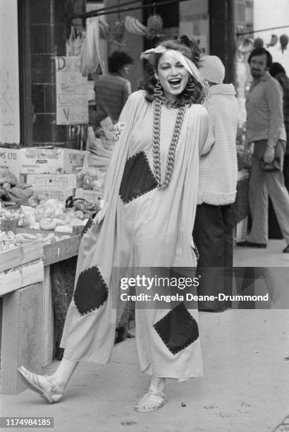 Fashion model wears a wide jumpsuit with patch inserts, an oversize chain necklace and jelly shoes as she walks through a street market, UK, 18th...