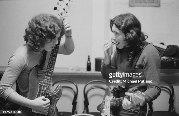 Irish singer and guitarist Rory Gallagher with his Fender Stratocaster strapped on, plays a harmonica backstage with bass guitarist Gerry McAvoy...