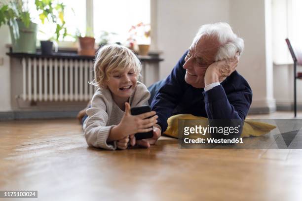 grandfather and grandson lying on the floor at home using a smartphone - seniors having fun with grandson stockfoto's en -beelden
