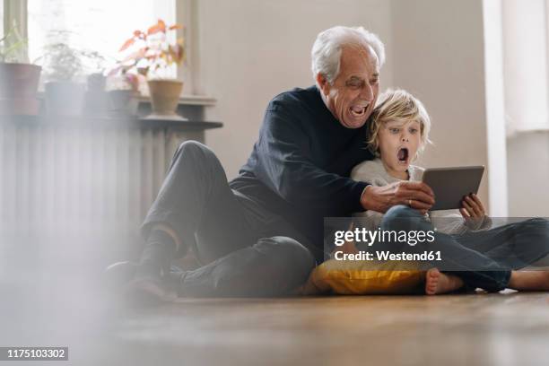 screaming grandfather and grandson sitting on the floor at home using a tablet - familie unterwegs stock-fotos und bilder