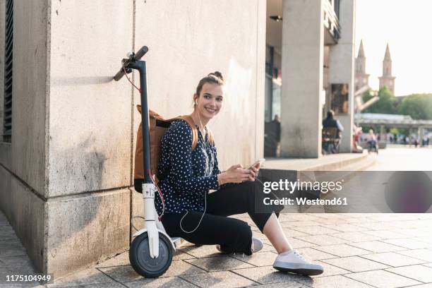 smiling young woman with electric scooter, earphones and cell phone having a break in the city - mobility scooter photos et images de collection